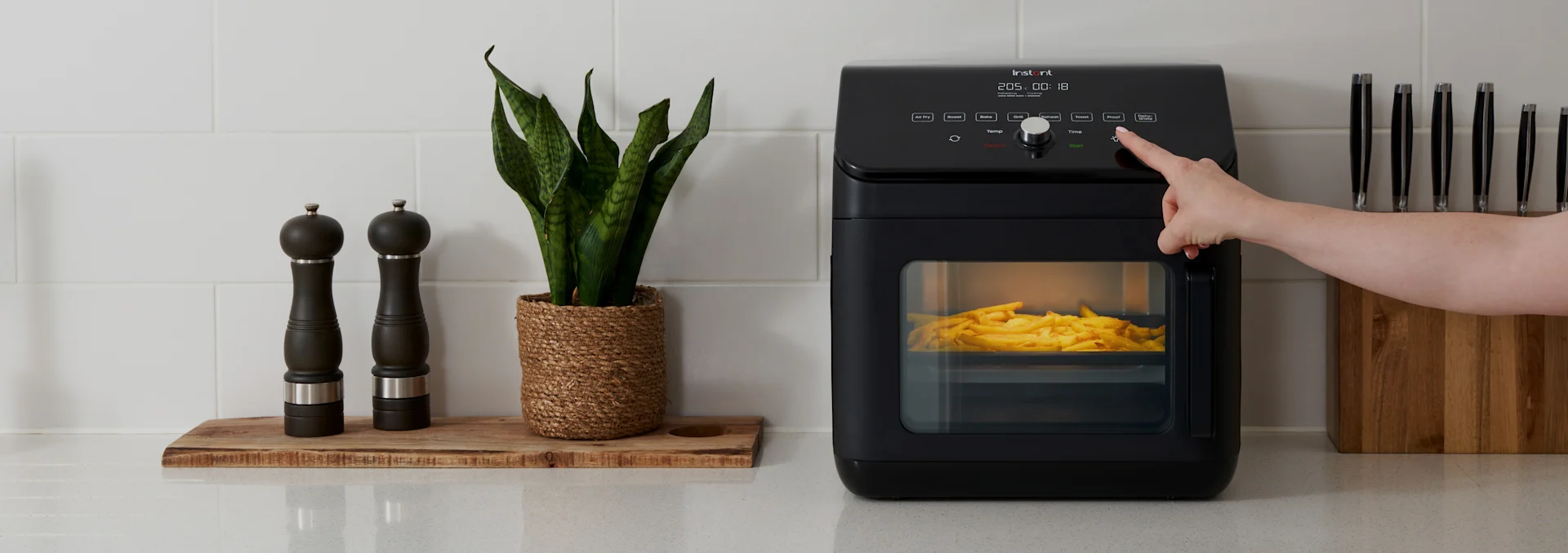 Käsi painaa nappia mustassa ilmakeittimessä, jossa on ranskalaisia, maustemyllyjen, kasvin ja veitsitelineen vieressä keittiötasolla.